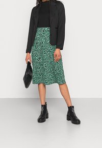 Blazer ajustado negro sobre una blusa negra de canalé, combinado con una falda verde floral y botas negras hasta el tobillo. Modelo sosteniendo un bolso negro.