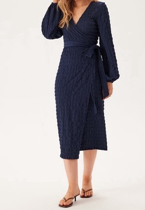 Navy blue wrap dress featuring long sleeves, textured fabric, and a tied waist. Midi length, with a subtle pattern and V-neck design.