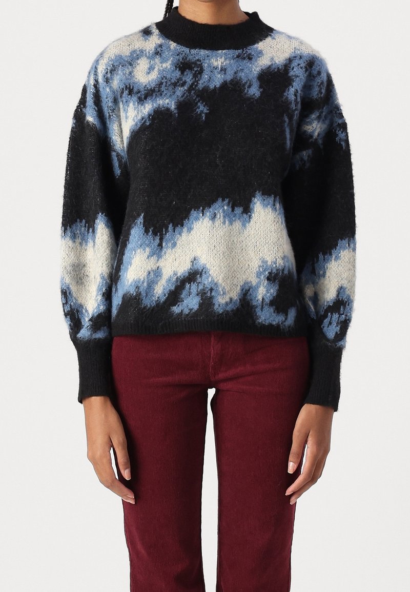 Pull en maille duveteuse noir, bleu et blanc avec un motif abstrait, porté avec un pantalon en velours côtelé rouge foncé, visible du cou à la moitié de la cuisse.