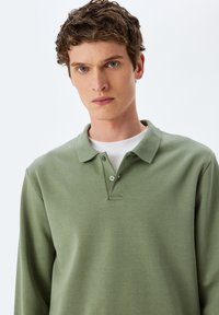 Langarm-Polohemd aus schimmerndem grünem Stoff mit weicher Textur. Verfügt über einen klassischen Kragen, eine Zwei-Knopf-Leiste und ein weißes Rundhalsunterhemd.