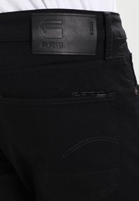 Jeans de mezclilla negra con un parche de cuero texturizado con logo grabado, costuras visibles y un diseño de bolsillo trasero con un acento curvado.