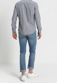 Jasnoszara koszula na guziki z długimi rękawami, w połączeniu z podwiniętymi jasnoniebieskimi jeansami i białymi trampkami, widziana od tyłu.