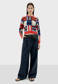 Un pull en tricot coloré avec des motifs géométriques rouges, bleus et bruns, associé à un pantalon large bleu marine et un sac banane noir.
