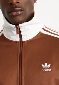Smeđa jakna na patentni zatvarač s bijelim ovratnikom i tri bijele pruge na ramenima, koja ima mali bijeli Adidas logo na prsima.