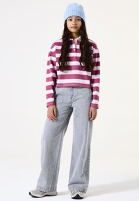 Gestreiftes, kurzes Polo in Burgunder, Pink und Weiß, kombiniert mit hellgrauen, weiten Jeans. Eine blaue Strickmütze rundet das Outfit ab.