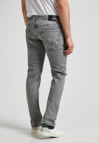 Uomo che indossa jeans grigi e sneakers bianche con dettagli verdi, in piedi contro uno sfondo chiaro e uniforme.