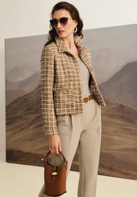 Vrouw in een tweed jasje, beige broek, grote gouden oorbellen en zonnebril, met een bruine handtas tegen een bergachtergrond.