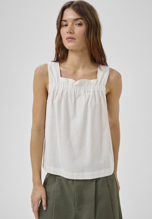 Kobieta o prostych brązowych włosach, ubrana w białą, bezrękawną, marszczoną bluzkę i oliwkowozielone spodnie, stojąca na tle jednolitego szarego tła.