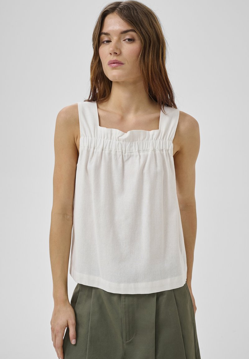 Femme aux cheveux bruns raides portant un haut blanc sans manches froncé et un pantalon vert olive, debout devant un fond gris uni.