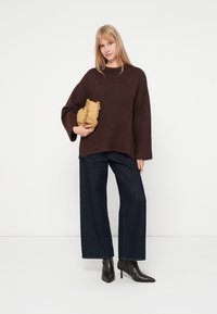 Pulover maro tricotat cu detalii în relief, asortat cu pantaloni largi din denim închis la culoare și cizme negre cu vârf ascuțit. Ține în mână o geantă plic texturată galbenă.