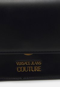 Mustan tekonahkainen laukku, jossa on kiiltävä pinta. Esittelee näkyvästi kultaisen logotekstin "VERSACE JEANS COUTURE". Siisti, minimalistinen muotoilu.