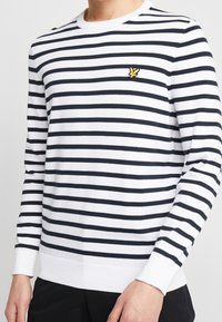 Maglione a righe bianche e blu navy con maniche lunghe, scollatura rotonda, polsini e orlo a coste, caratterizzato da un piccolo logo giallo sul petto.