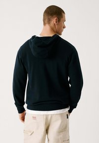 Marinblå hoodie med justerbar huva, långa ärmar, ribbade muddar och nederkant. Bärs med en lager-vit skjorta som har en slät yta.