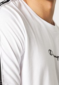 Hvid kortærmet T-shirt med rund hals, med et sort Champion logo og stribede sorte detaljer på skuldrene. Blødt stof.