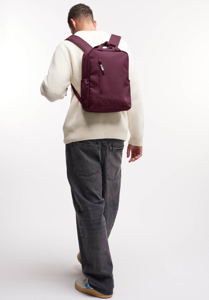 Sac à dos en nylon bordeaux de forme rectangulaire, avec une poche zippée à l'avant et des bretelles réglables, porté sur un pull crème.