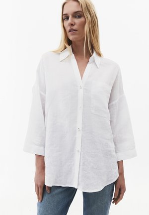 Mujer rubia que lleva una camisa blanca holgada con botones y un bolsillo en el pecho, y vaqueros azul claro, sobre un fondo liso.