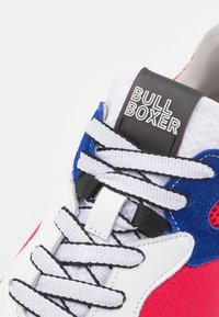 Weiße und blaue Sneaker mit roten Akzenten, ausgestattet mit Mesh-Material, strukturierten Stoffüberzügen, einem schwarzen Logo-Patch und flachen weißen Schnürsenkeln.