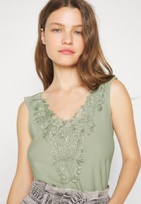 Top verde sem mangas com decote em V, apresentando bordados florais intricados e detalhes recortados ao redor do decote. Tecido suave e liso.