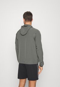 Chaqueta ligera con capucha de color gris, mangas largas, puños elásticos y dobladillo recto. Colocada sobre pantalones cortos negros, con un diseño sencillo.