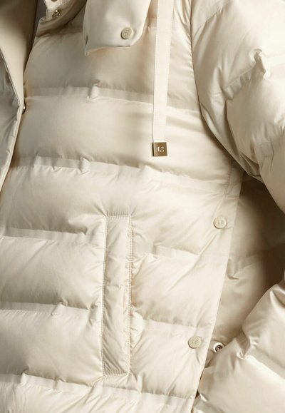 Beige pufferjack met een slanke, gewatteerde uitstraling. Bevat knoopsluitingen aan de voorkant, zijzakken en een capuchon met trekkoord voor een verstelbare pasvorm.