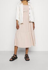 Světlounce růžové midi šaty s nařaseným živůtkem, vzdušnou sukni s volánky a velkou kulatou sponou na pásku. Stylizované s bílou džínovou bundou a černými sandály.