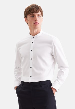 Jeune homme portant une chemise blanche à col mandarin avec des boutons noirs et un pantalon noir, mains dans les poches, regardant légèrement vers la droite.