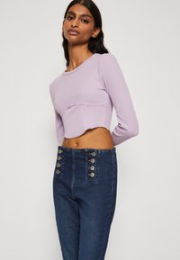 Top de corto de manga larga con canalé en color lavanda, con un dobladillo redondeado, combinado con pantalones de mezclilla oscuros de tiro alto que presentan detalles de botones plateados.
