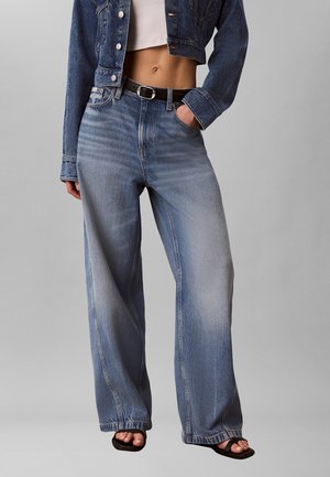 Osoba nosząca krótki jeansowy kurtkę, biały krótki top, wysokie spodnie dżinsowe o szerokich nogawkach w kolorze wyblakłego niebieskiego, czarny pasek i czarne sandały z odkrytymi palcami.