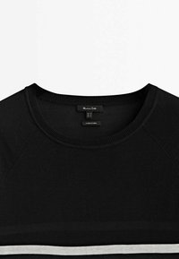 Pull noir à col rond avec col côtelé et bande blanche horizontale près du bord inférieur, montrant une étiquette de taille M et une étiquette "Fabriqué en Chine".