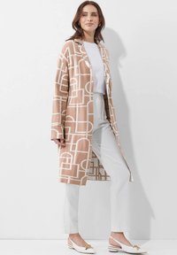 Beige lange cardigan met witte geometrische patronen, gelaagd over een witte top, gecombineerd met een lichte broek en beige slingback schoenen.