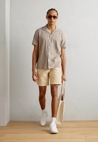 Une chemise beige à boutons, un short clair, des baskets blanches, portant un sac fourre-tout beige, avec des lunettes de soleil, marchant devant un fond neutre.