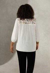 Vit blus med en genomskinlig spetsbakpanel, mjuka ärmar och en flytande passform. Kombineras med svarta byxor som visar ett strukturerat tyg.