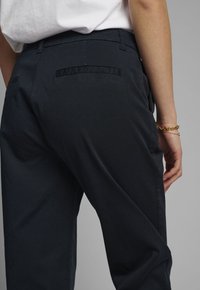 Pantalon noir en tissu de coton, présentant une coupe droite, une texture lisse et deux poches arrière avec des détails cousus.