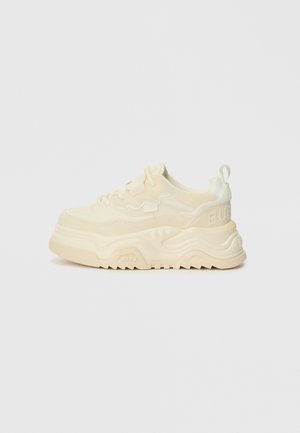 Ténis robustos e brancos cremosos com camurça texturizada e acabamentos lisos, apresentando uma sola grossa, fecho de atacador e um logótipo proeminente no calcanhar.