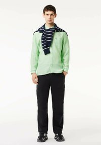 Camisa de botones a rayas verdes y blancas, suéter a rayas marinas alrededor de los hombros, pantalones cargo negros y zapatillas negras.