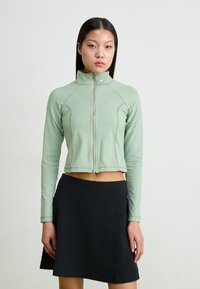 Minta zaļā īsā jaka ar rāvējslēdzēju priekšpusē, garām piedurknēm un kontrastējošiem šuvēm, savienota ar melnu ribotu svārku.