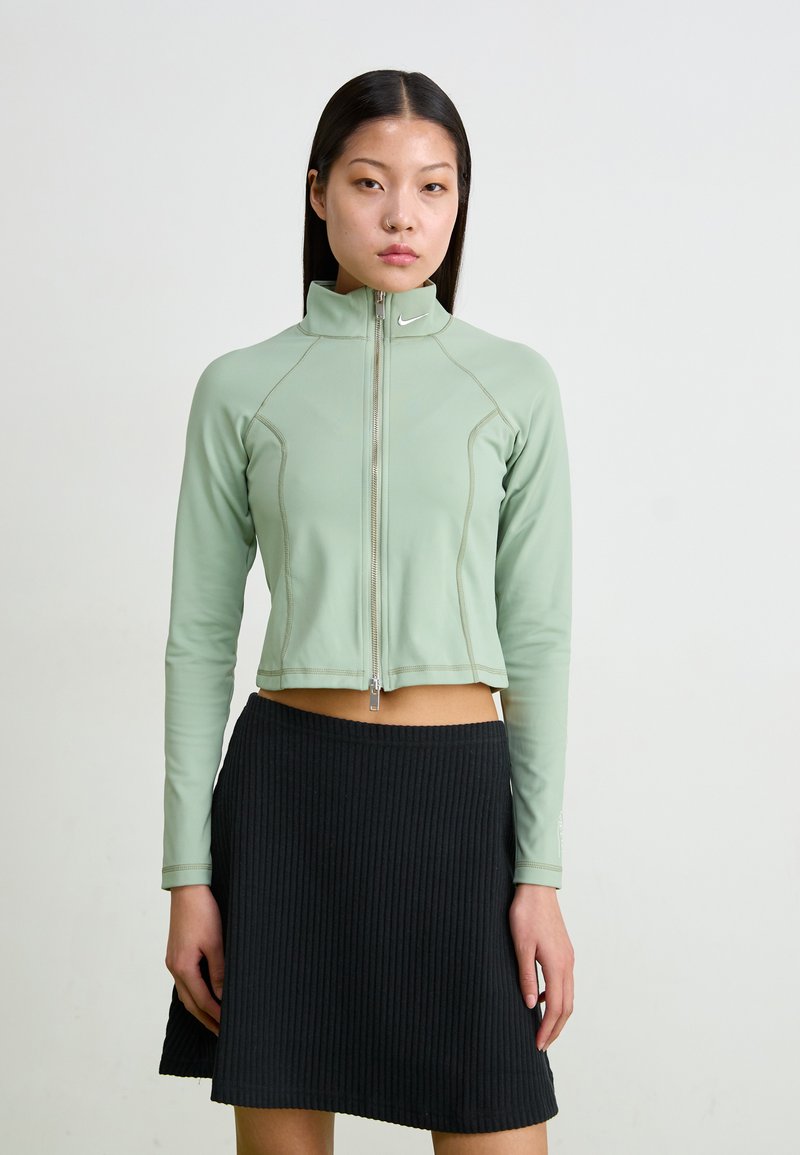 Minta zaļā īsā jaka ar rāvējslēdzēju priekšpusē, garām piedurknēm un kontrastējošiem šuvēm, savienota ar melnu ribotu svārku.