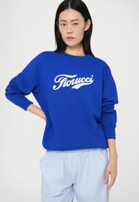 Sieviete ar taisniem melniem matiem, tērpta zilajā Fiorucci sporta džemperī un gaiši zilās svītrainās biksēs, stāv ar vienu roku uz gurniem.