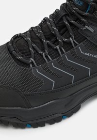 Scarpe alte impermeabili nere con mesh testurizzato e sovrapposizioni sintetiche, con accenti grigi e dettagli blu sulla suola.