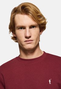 Burgundy sweatshirt med ribbad rund hals, mjuk textur och vit broderad logotyp på vänster bröst. Klassisk design, avslappnad passform.