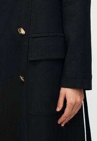 Κοντινό πλάνο μανικιού μαύρου παλτού, πλευρική τσέπη με καπάκι και δύο χρυσά κουμπιά με ανάγλυφο κείμενο κοντά σε ένα χαλαρό χέρι.