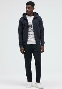 Navy zip-up hoodie with drawstring, layered over a white graphic T-shirt. Paired with dark trousers and white sneakers.