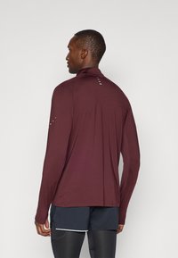 Cămașă sport cu mâneci lungi într-o nuanță profundă de burgund, cu un gât înalt, detalii fine pe cusătura din spate și accente reflective pe umeri.