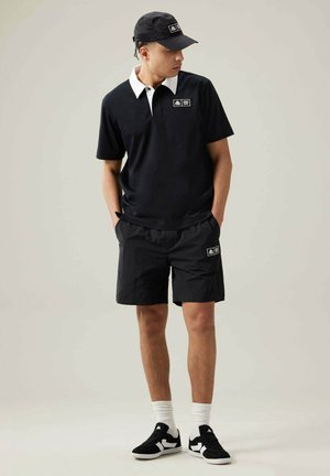 Hombre con camiseta polo negra con cuello blanco, pantalones cortos negros, gorra negra, calcetines blancos y zapatillas negras, de pie con las manos en los bolsillos.