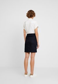 Jupe en denim bleu foncé avec une coupe ajustée, longueur mi-cuisse et passants de ceinture. Associée à une chemise blanche à manches courtes et des baskets blanches.