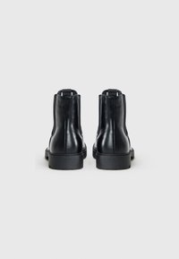 Botas de cuero negras hasta el tobillo con una textura suave, puntera redonda y un tacón bajo y grueso. Presenta una lengüeta en la parte trasera para facilitar su uso.