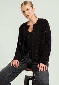 Cardigan nero con texture, scollo rotondo, bottoni frontali e sottili accenti glitterati. Abbinato a jeans grigio scuro, seduto su uno sgabello.