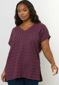 Kobieta ubrana w teksturowaną fioletową bluzkę z krótkim rękawem w kształcie litery V z bocznymi rozcięciami, zestawioną z ciemnoniebieskimi spodniami, na jednolitym białym tle.