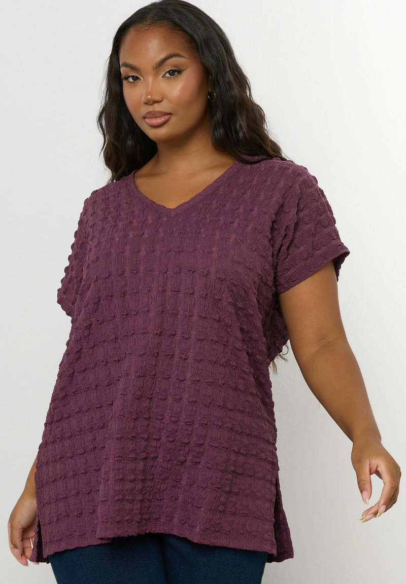 Kobieta ubrana w teksturowaną fioletową bluzkę z krótkim rękawem w kształcie litery V z bocznymi rozcięciami, zestawioną z ciemnoniebieskimi spodniami, na jednolitym białym tle.