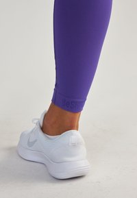 Leggings de sport violets au design ajusté, dotés d'un détail logo au niveau de la cheville, associés à des baskets en mesh blanches avec un accent gris.